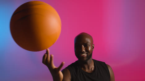 Studio-Portrait-Shot-Of-Male-Basketball-Player-Spinning-Ball-On-Finger-Against-Pink-And-Blue-Lit-Background
